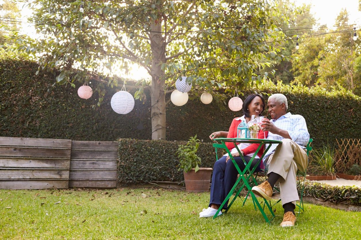 People Sitting At A Table In The Yard With Plants And Lights Hanging From The Tree