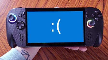 Hand holding ROG Xbox Ally X with blue screen and sideways sad face above woodgrain table.