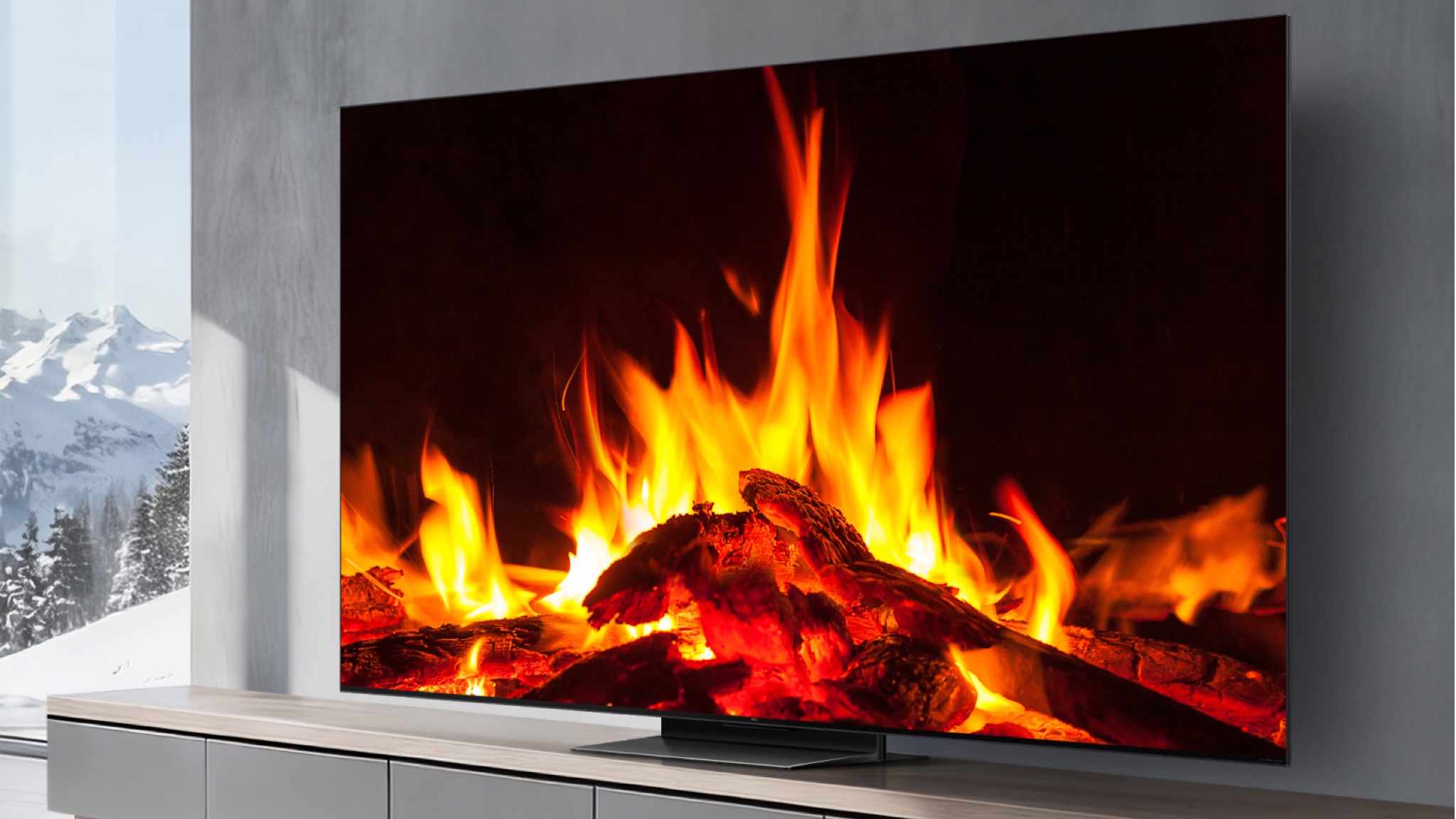 A TCL C7L TV showing a wood fire. It is in a grey decorated room with snowy mountains visible through a full height window to the left of the tv unit