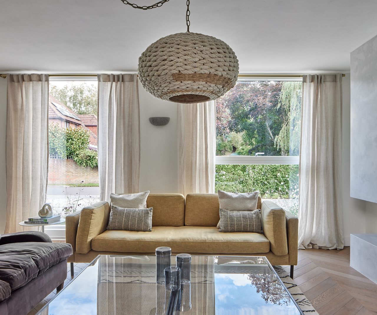 A soft yellow sofa with neatly arranged cushions with two large modern glazed windows behind. There is a central glass coffee table over which hangs a large lamp shade