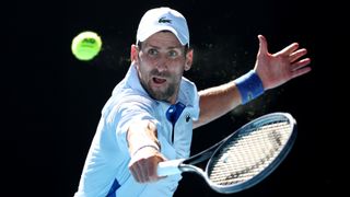 Novak Djokovic of Serbia, wearing white baseball cap and shirt, plays a backhand at the Australian Open 2024 Grand Slam tennis tournament. 