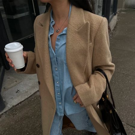 Sasha Mei wearing a camel coat, denim shirt, and jeans with a black tote bag. 