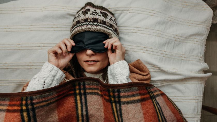A woman sleeping cozy in winter by layering up on bedding, wearing a hat, a sleep mask and snuggled under a tartan blanket.