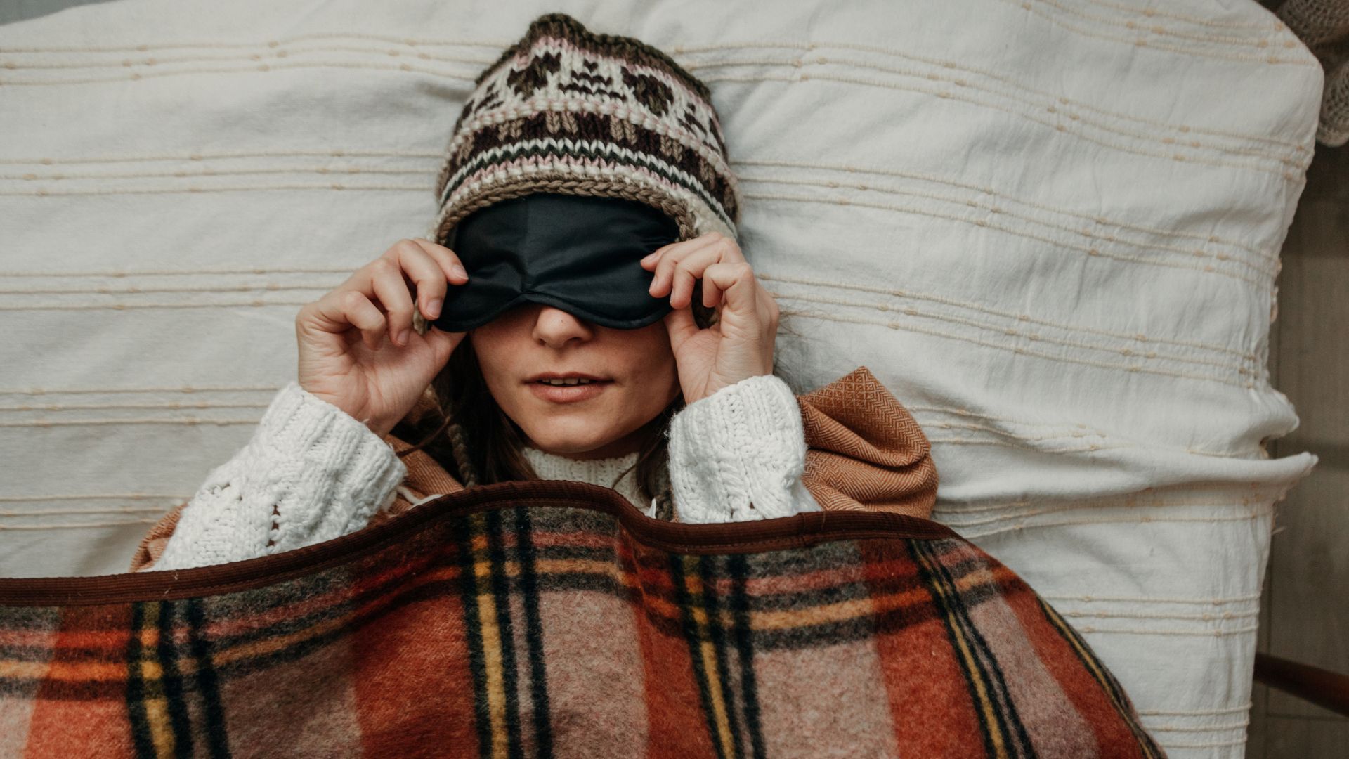 A woman sleeping cozy in winter by layering up on bedding, wearing a hat, a sleep mask and snuggled under a tartan blanket.