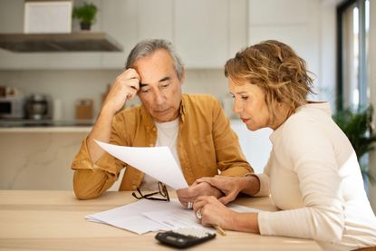 2R069T3 Concentrated senior spouses sitting at table full of papers and counting monthly expenses, kitchen interior