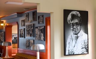 A large black & white photo of a black female is hung on the wall to the right. Through an opening to the left, we see black & white photographs of black female models hung on a light blue and white dotted wall.
