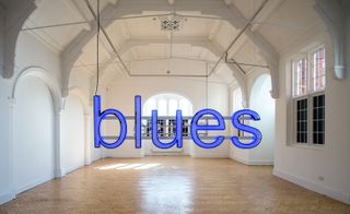 The word "blues" gently lit in blue, is suspended in a white room with an arched ceiling