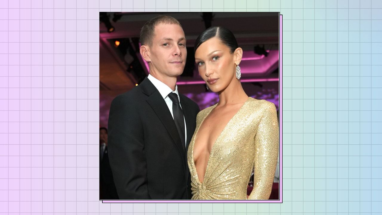 Marc Kalman and Bella Hadid, who is wearing a gold, v-neck dress, posing together the Golden Heart Awards 2022 Benefiting God&#039;s Love We Deliver at The Glasshouse on October 17, 2022 in New York City | on a purple, green and blue background