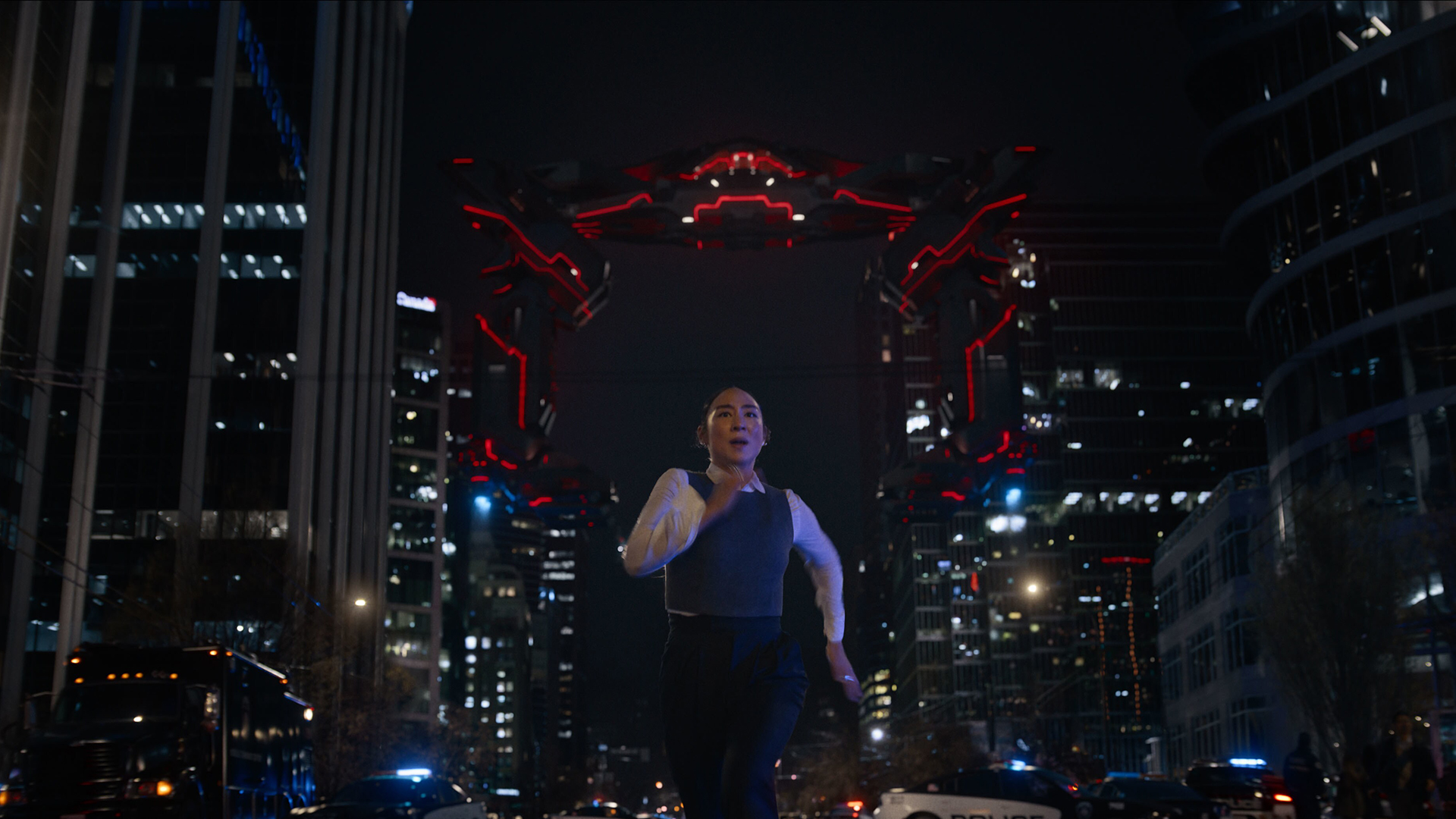 Still from the movie Tron: Ares. A woman is frantically running away from a menacing red and black futuristic drone monster. It is nighttime and dark as she runs between skyscrapers.