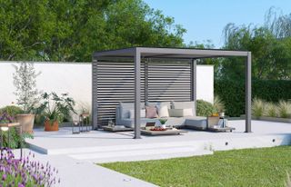 Louvered pergola with a roof, garden sofa and firepit, on a deck with potted plants and flowers, with trees in the background and lawn in the foreground