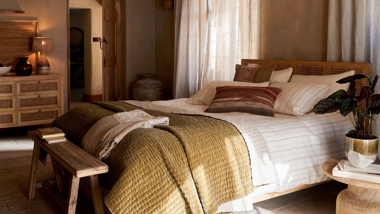 An earth toned bedroom with a yellow blanket draped over the bed, and stacks of throw pillows. A small wooden bench at the base of the bed.