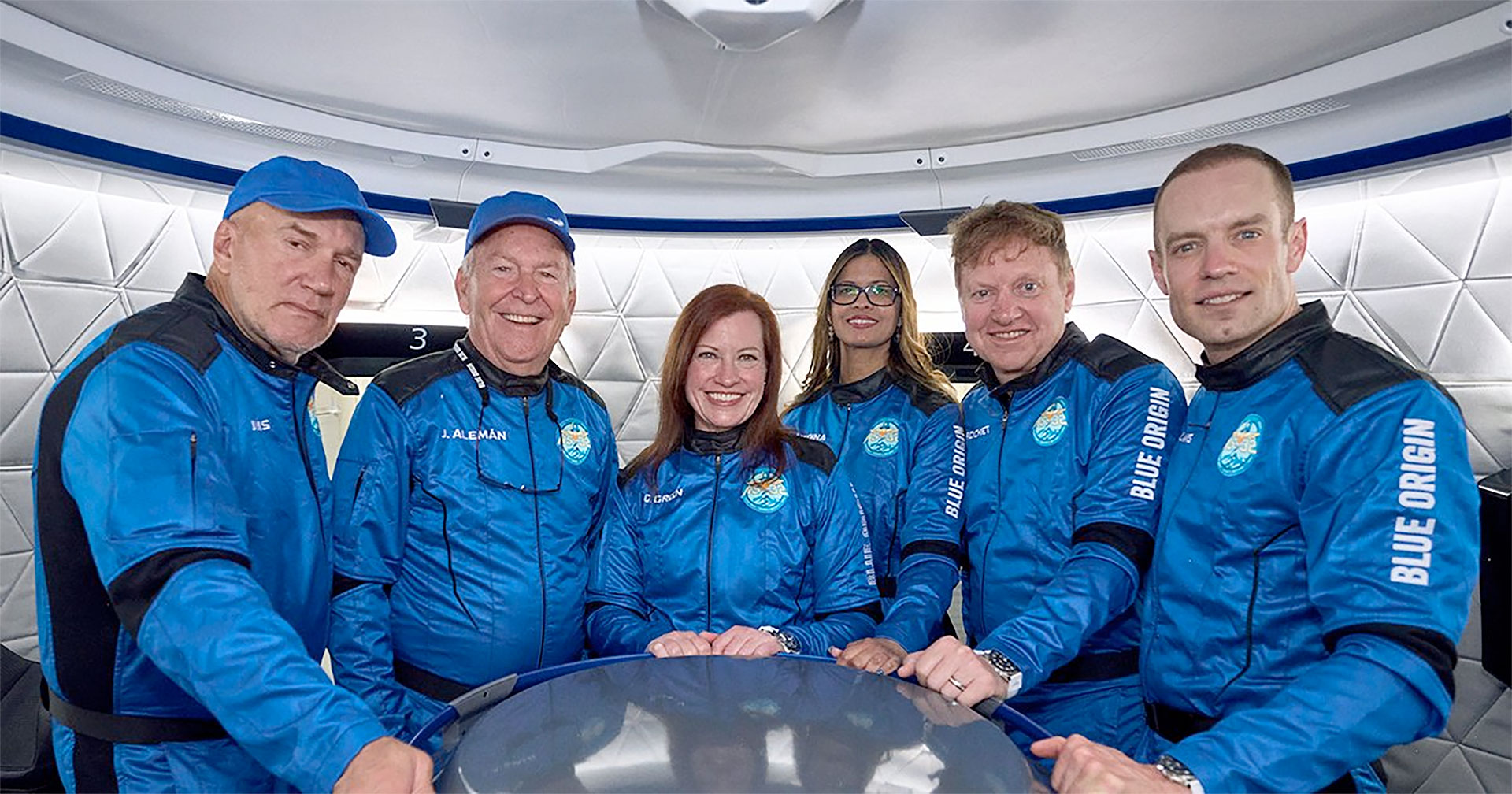four men and two women dressed in blue and black flight suits stand inside a mock space capsule