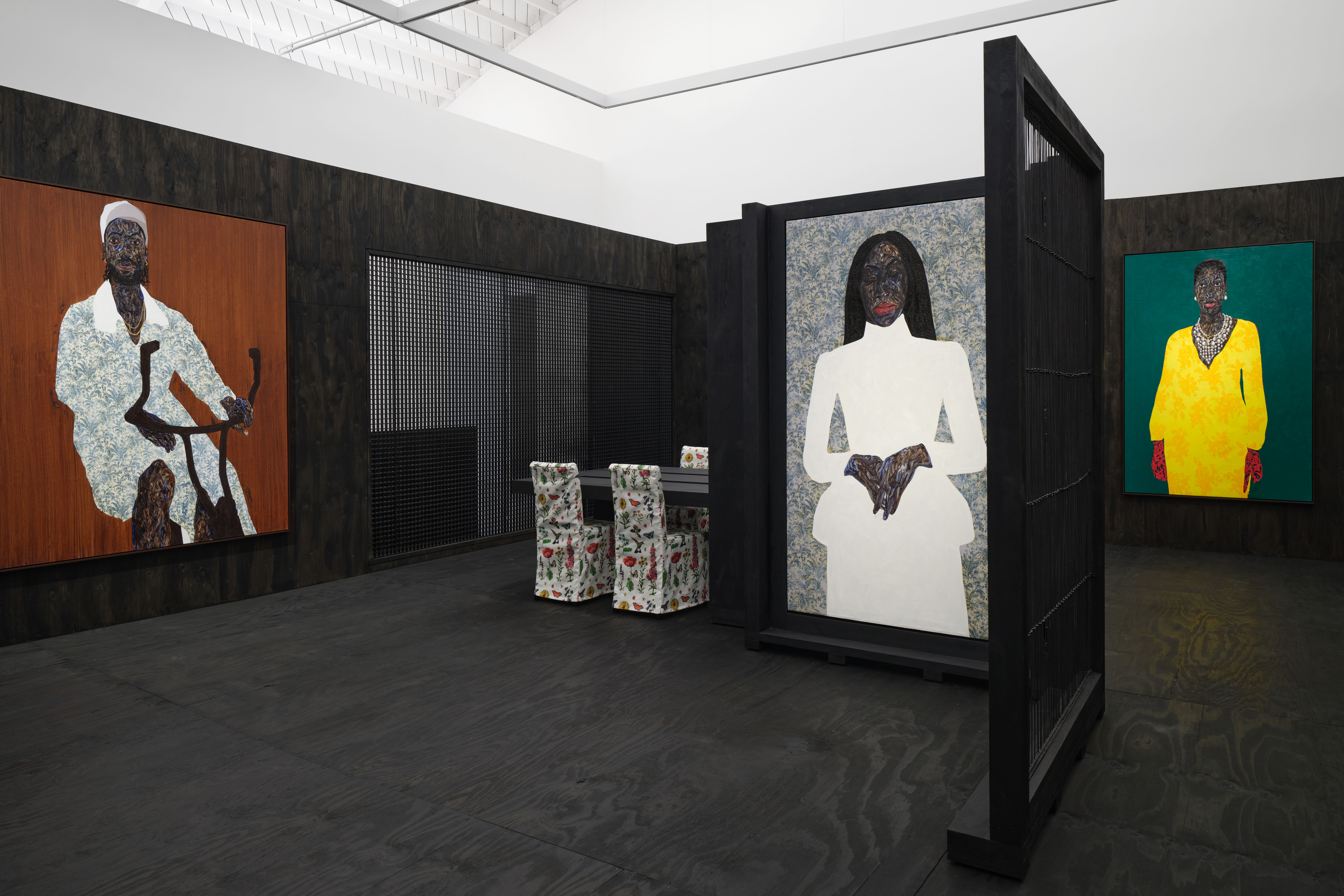 View of an art gallery installation with dark wood walls and multiple expressive portrait paintings. In the foreground, a large black metal structure with vertical bars frames additional paintings inside, while more portraits hang on the walls in the background.