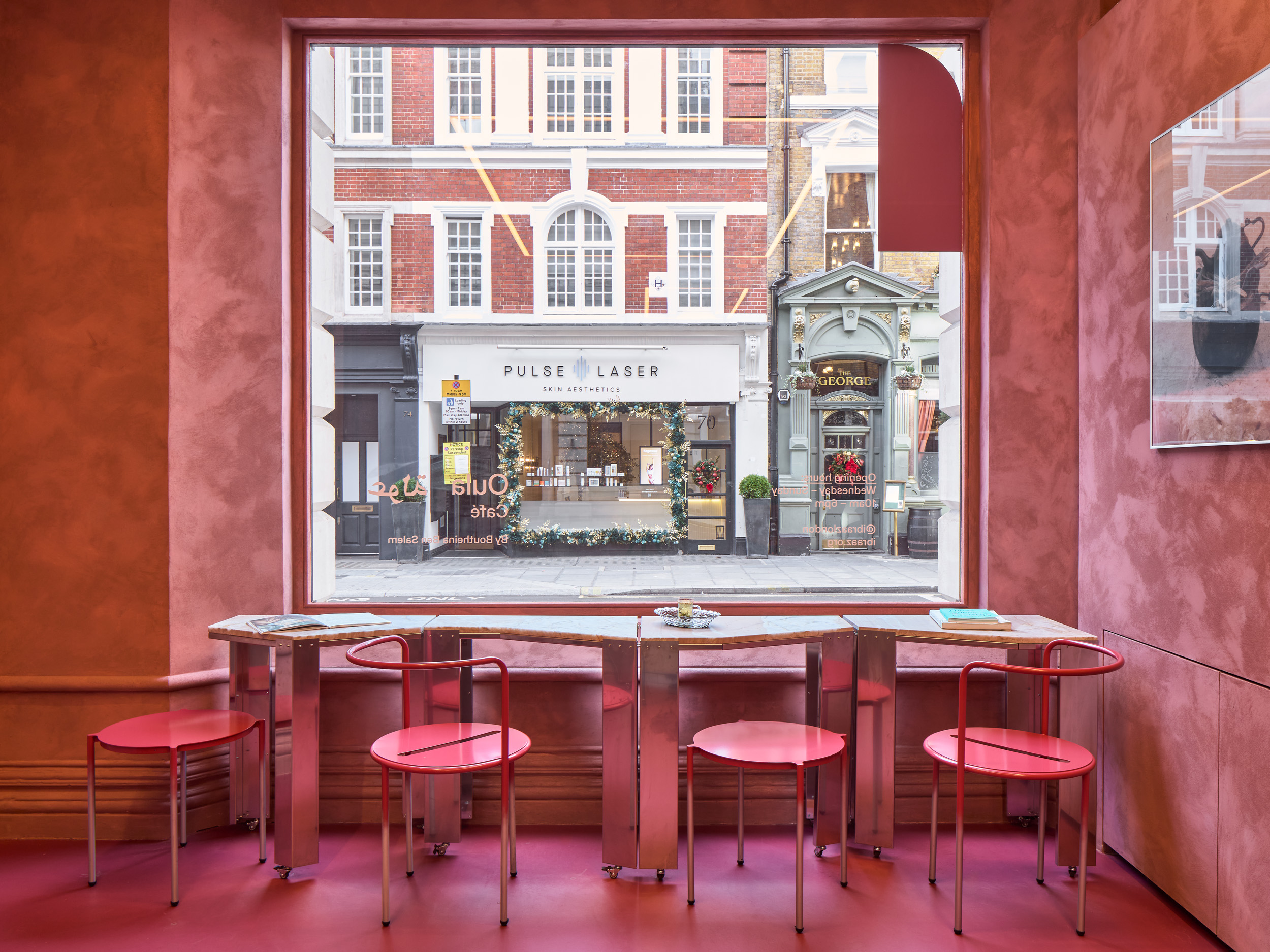 A pink-painted cafe with bright pink and chrome chairs, marble-topped sculptural tables with chrome legs, and lots of deli products and artworks styled on its shelves.