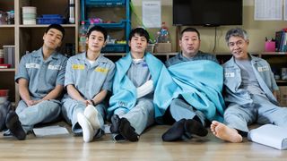 Five men wearing gray prison uniforms sit in a row, some wearing blankets, in 'Prison Playbook.'