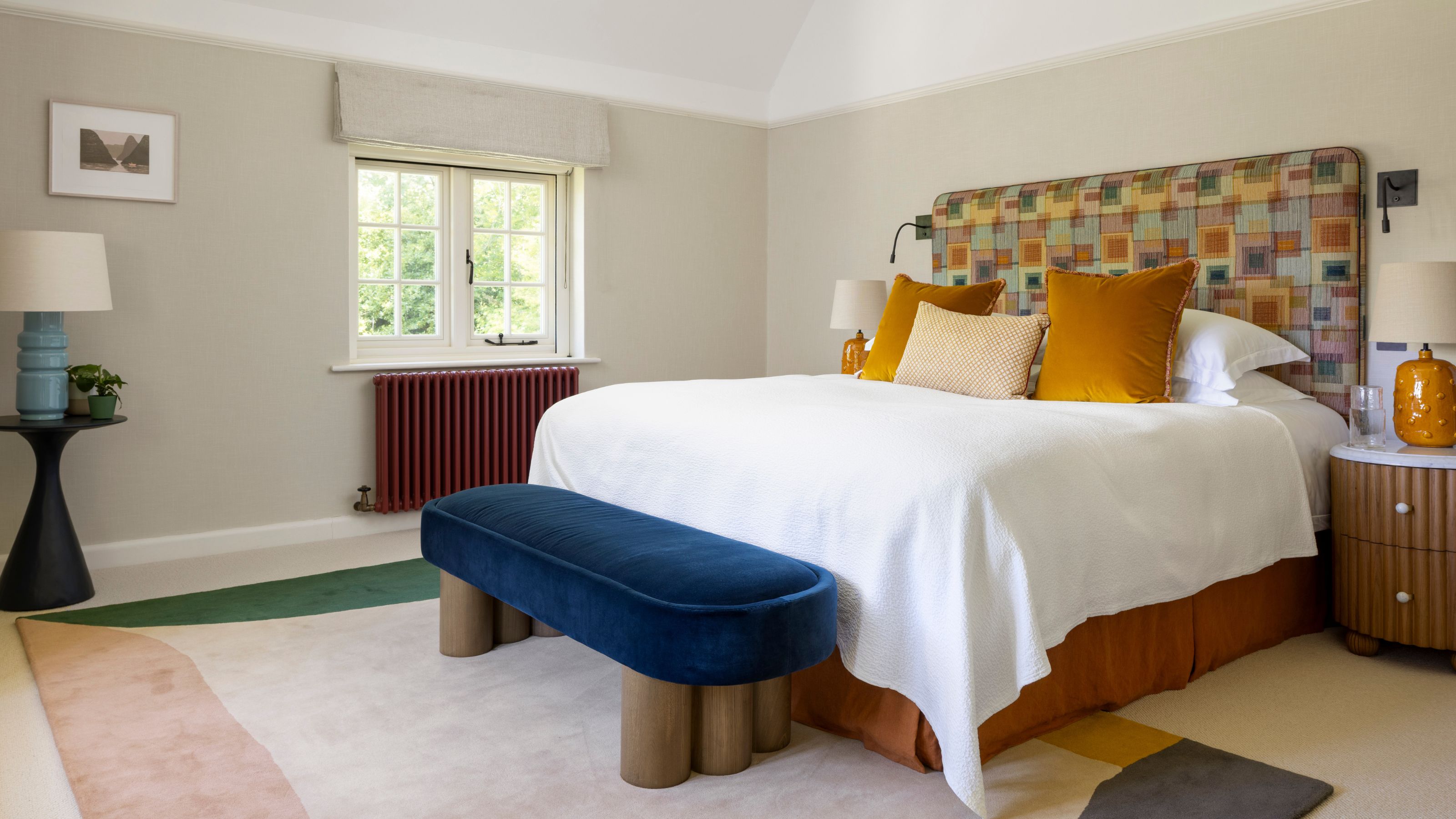 a neutral primary bedroom with an abstract rug, upholstered headboard, mustard pillows, navy velvet end of bed bench and a burgundy painted radiator