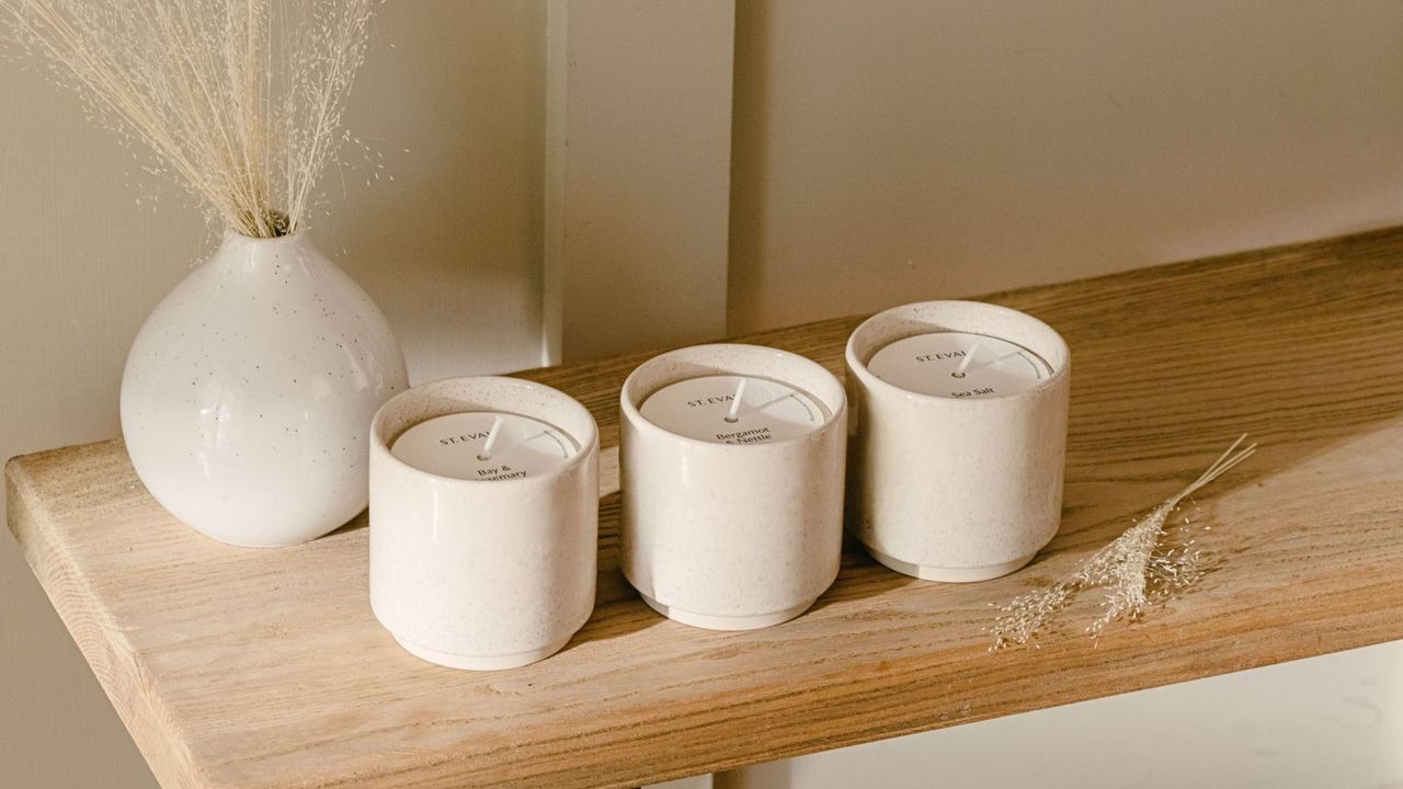 Three candles in neutral colored pots on a wooden sideboard beside a vase of dried stems