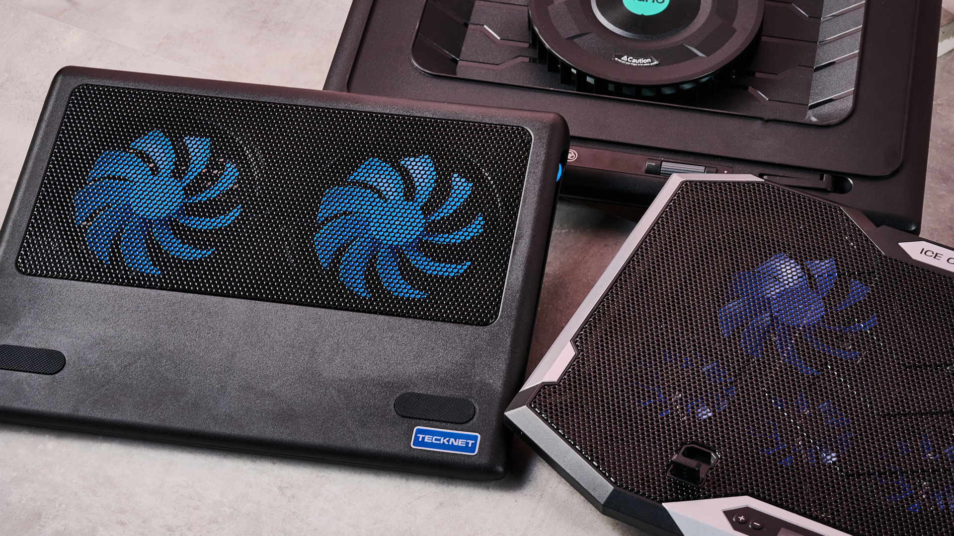 A closeup of the Llano RGB Laptop Cooling Pad, ICE COOREL RGB Laptop Cooling Pad and Tecknet N8 on a gray marbled desktop in front of a pink background.