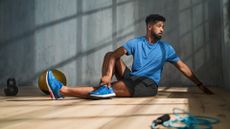 Young African American sportsman sitting on floor and doing stretching exercise indoors, workout training concept