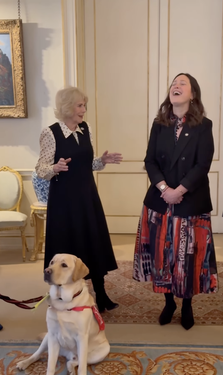 Queen Camilla talking to a woman and laughing with a dog sitting in front of them