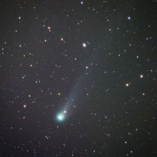 a green glowing nucleus of the comet is visible with a long tail against a star studded sky.