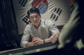 Wi Ha-jun as Kwon Jun-taek, sitting with his forearms resting on a table in front of a South Korean flag, in Gyeongseong Creature S1.