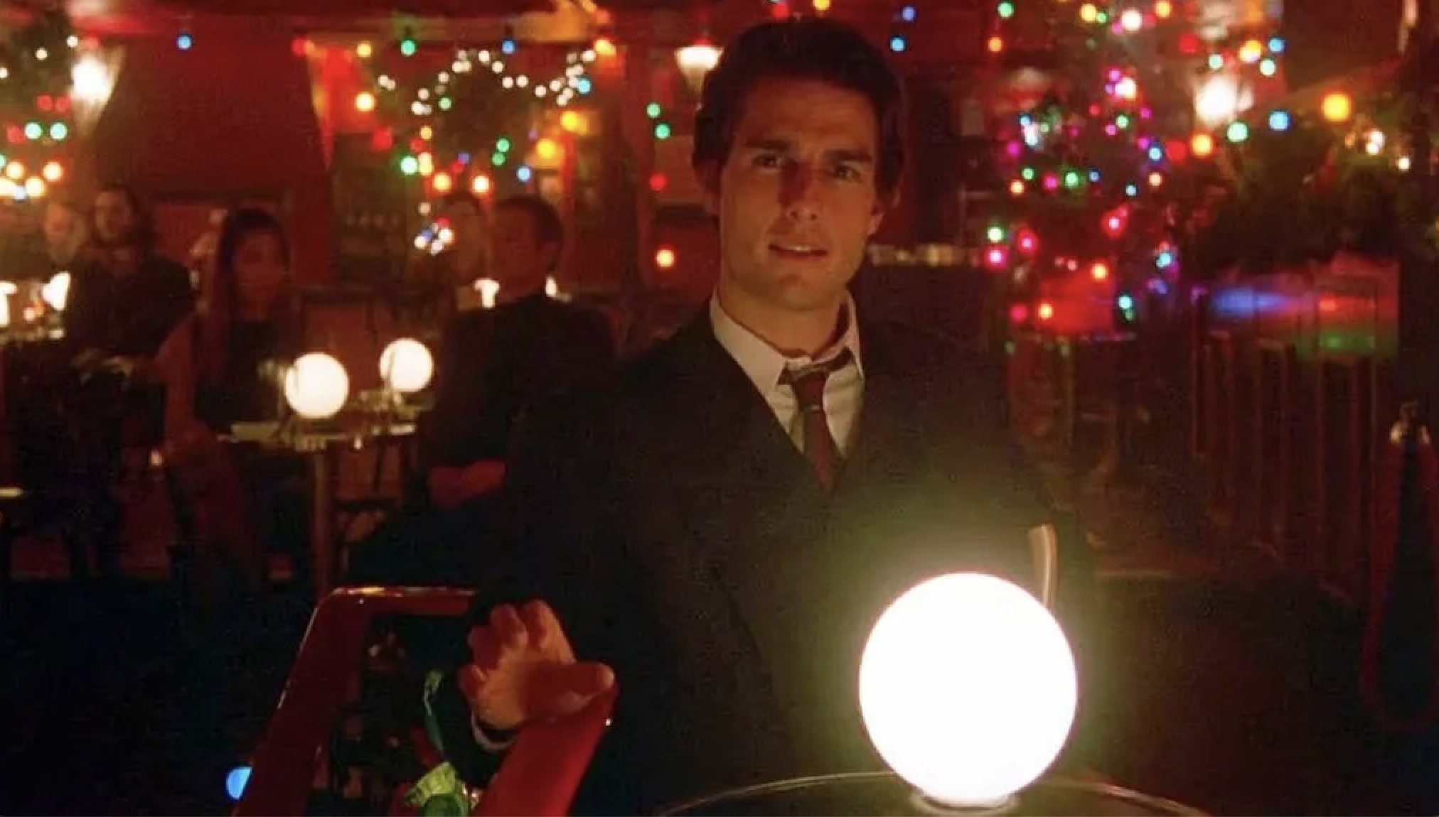 A man in a dark suit and loosened tie stands in a dimly lit room filled with colorful Christmas lights and glowing orb lamps.