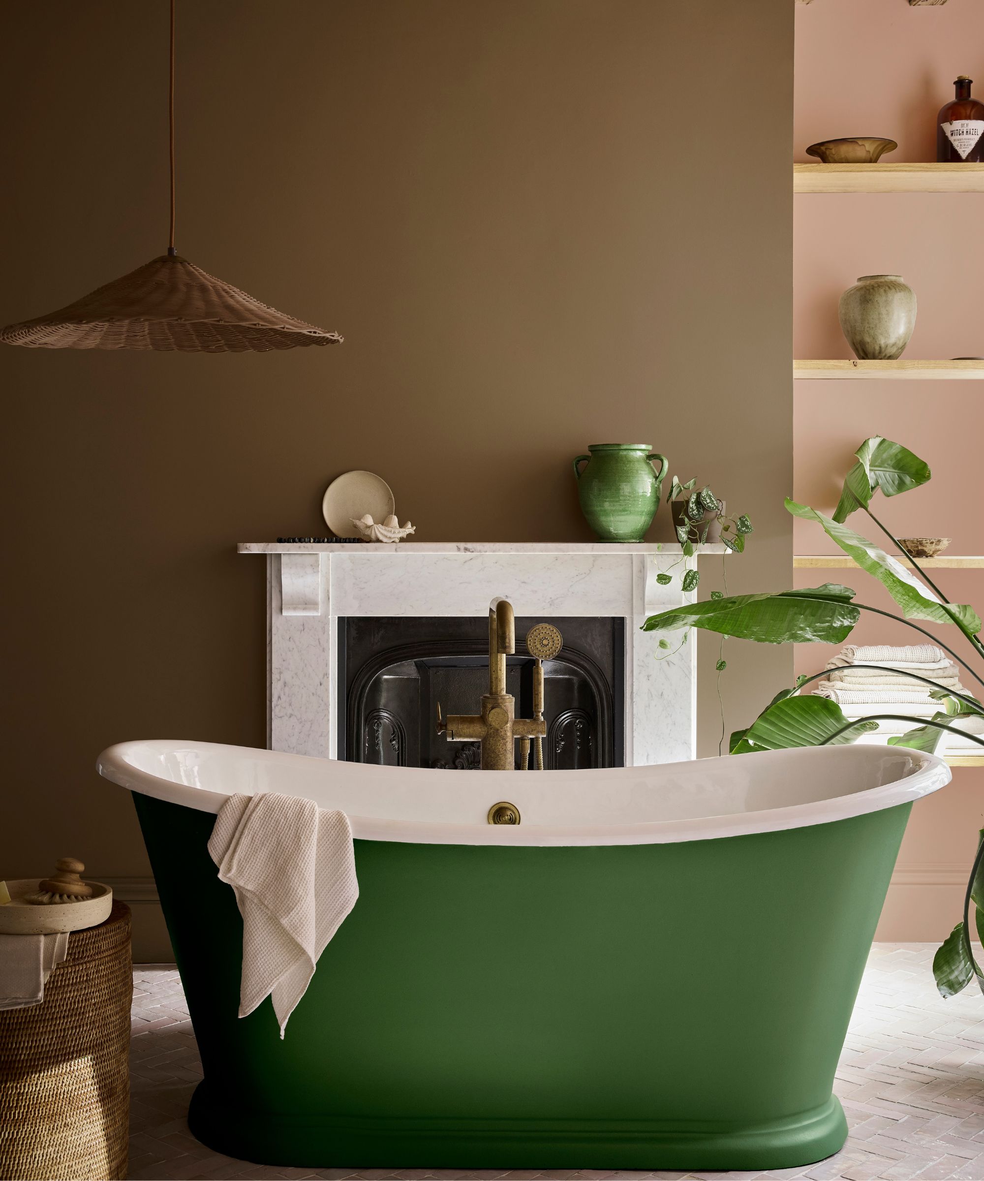 Bathroom with walls painted in Little Greene's Marle, and shelving painted in Mochi by Little Greene. A green freestanding tub sits in the centre, and a marble fireplace with ceramics on the mantel.