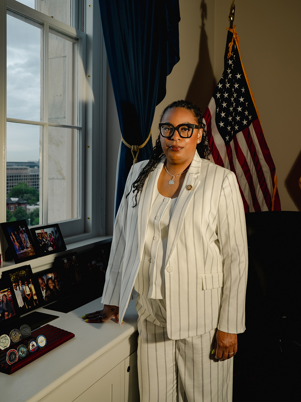 LaMonica McIver stands in her congressional office wearing a white pinstripe suit. An American flag is behind her; family photographs and challenge coins line the windowsill to her left.