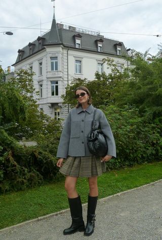 A woman wearing a plaid pleated miniskirt with a gray peacoat and black moto boots.