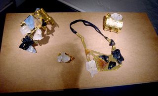 Four jewellery pieces displayed on a table. The left two are unrecognisable. The two on the right includes a neckless with brass hinge style connectors and emerald-like stones. The second right piece is a brass-like wrist item.