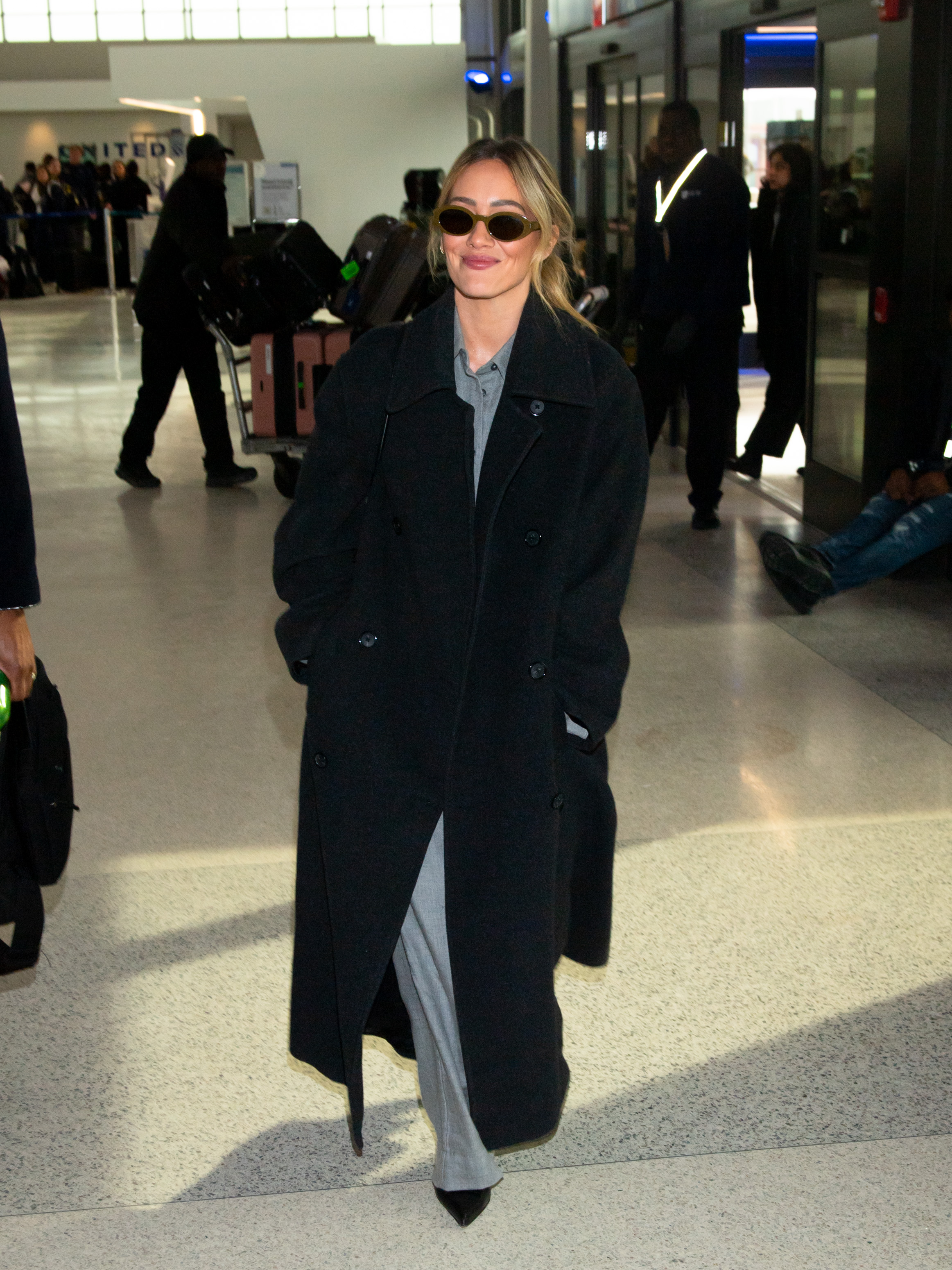 Hilary Duff wears a black double breasted coat, grey button down, grey trousers, pointy toe boots and brown oval sunglasses at the airport.