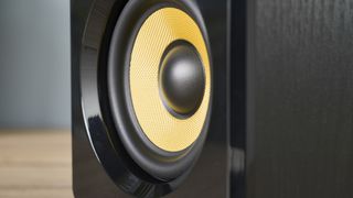 a pair of black wood speakers with a yellow kevlar cone photographed on a wood desk with a computer and blue wall and a plant in shot