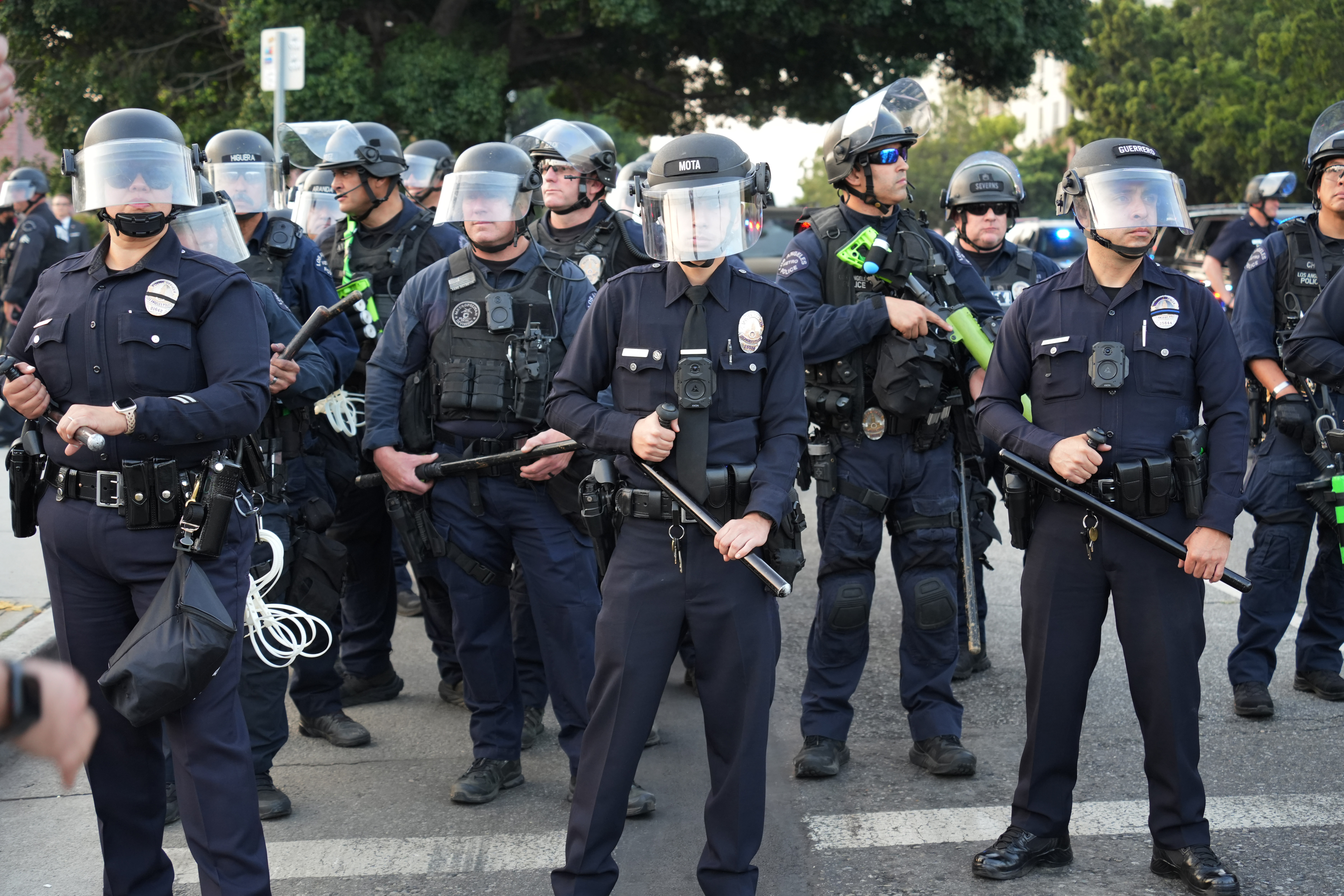 A line of police officers in riot gear block a city street.