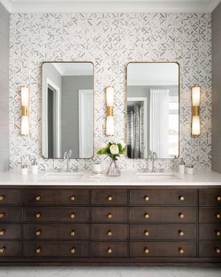 bathroom with grey and white patterned tiles, dark wood cabinetry, twin sinks, two mirrors, wall lights, brass hardware,