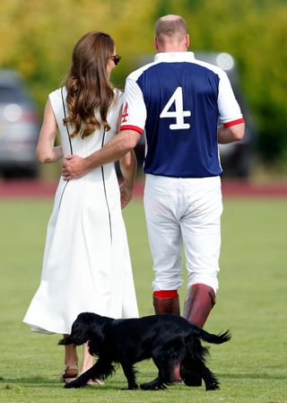 Princess Kate wearing a white dress and Prince William wearing white pants and a polo jersey walking away with their back to the camera and their black dog running behind them