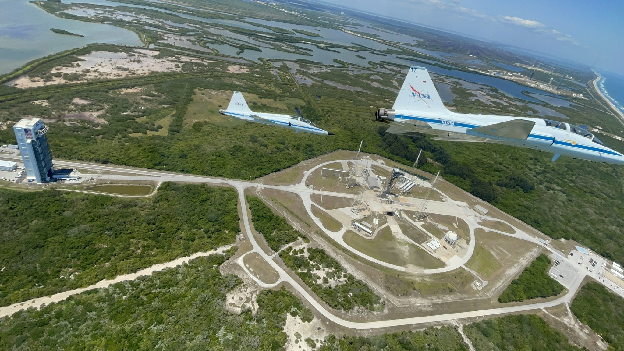 two jets over a launch pad. swamp is in the background. at left is a tall building