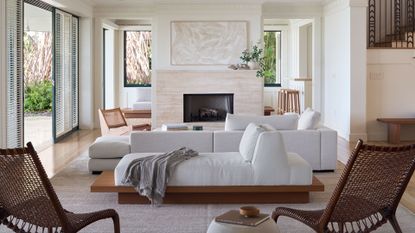 A bright, airy, white living room with a stone fireplace, a minimalist painting, a vase of green stems, a sectional white sofa, a chaise lounger, a pair of rattan chairs, a side table with a book and a bowl on top