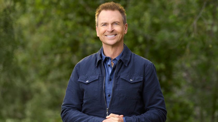 Phil Keoghan stands in front of trees in a dark blue shirt and jacket The Amazing Race Season 37