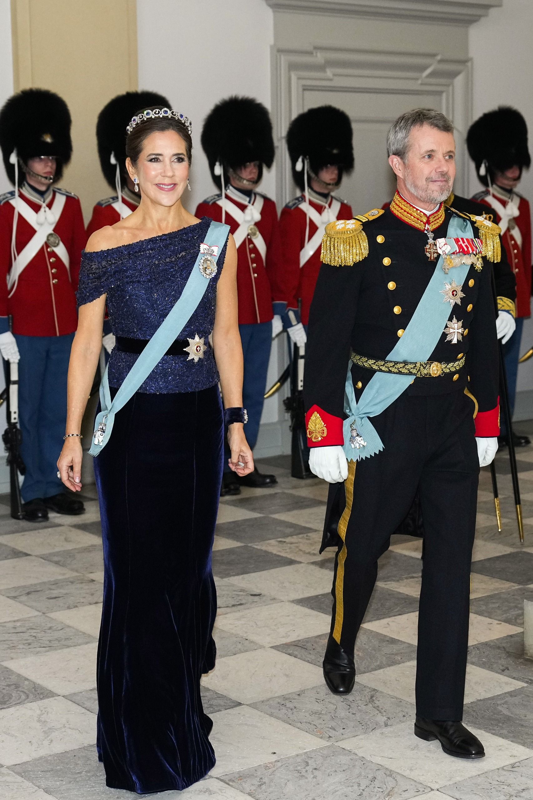 Queen Mary wearing a navy dress and tiara walking next to King Frederik in front of royal guards