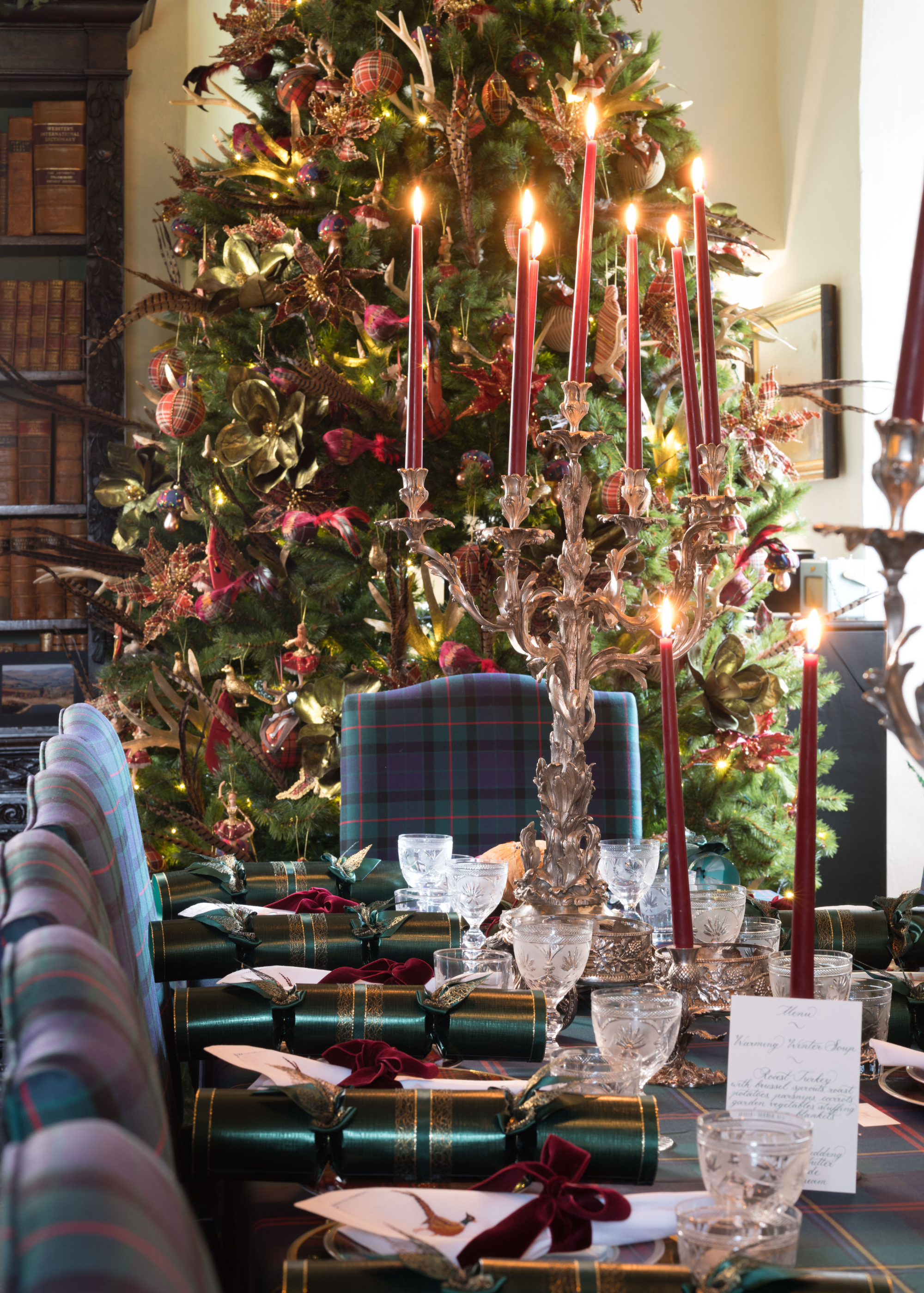 A dining area with a table set up with plates, glassware, long red candles and a large chrome candle holder. There is also a view of a well lit Christmas tree in the background.