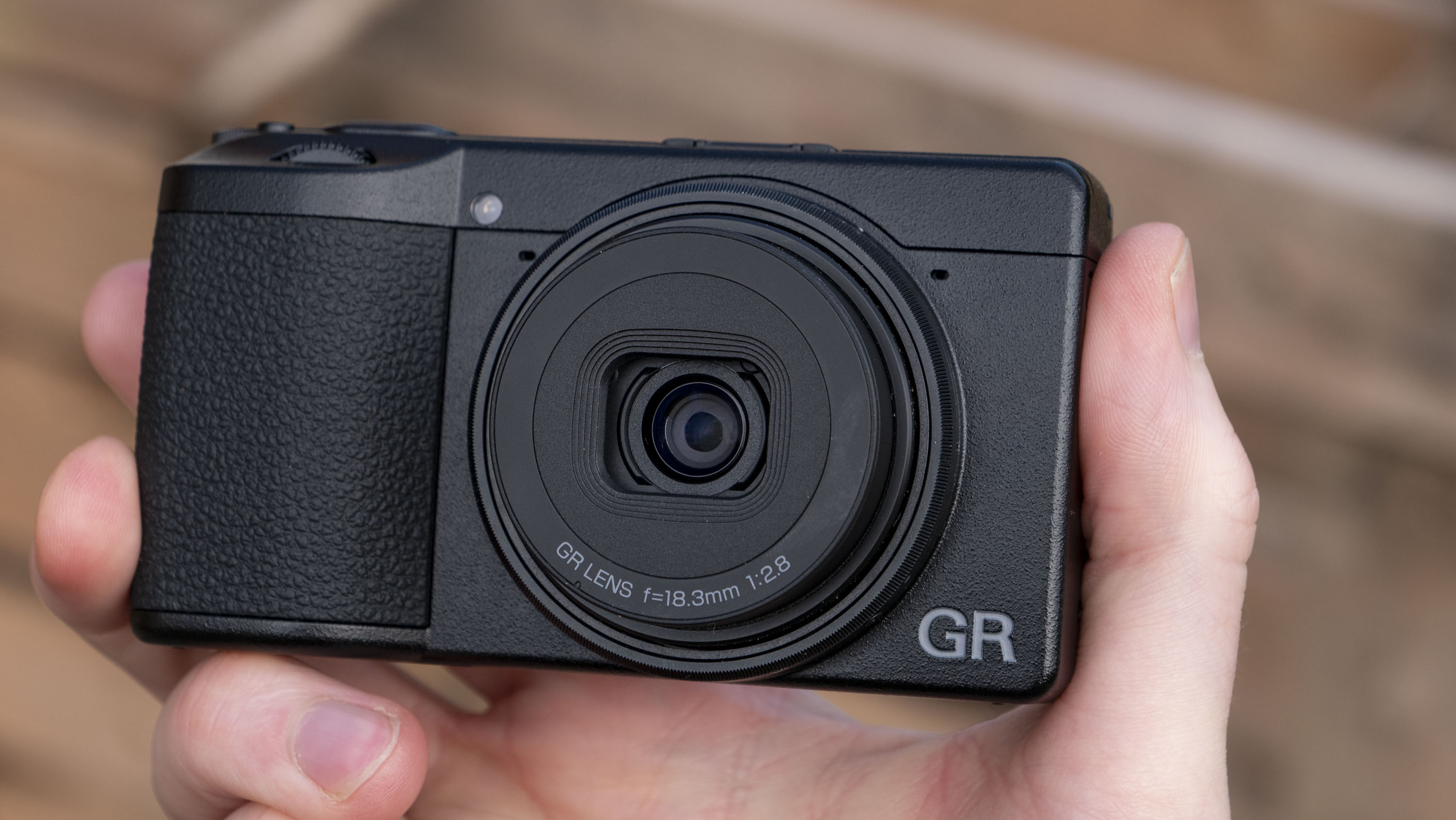 Ricoh GR IV compact camera in user's hand with wooden slats in the background
