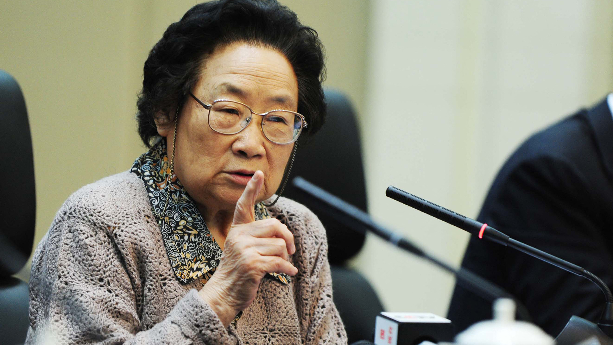 An Asian woman with short curly hair and glasses speaks into a microphone