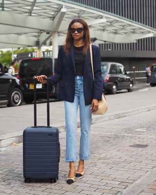 Woman traveling, wearing jeans, blazer, and blue carry-on suitcase at airport