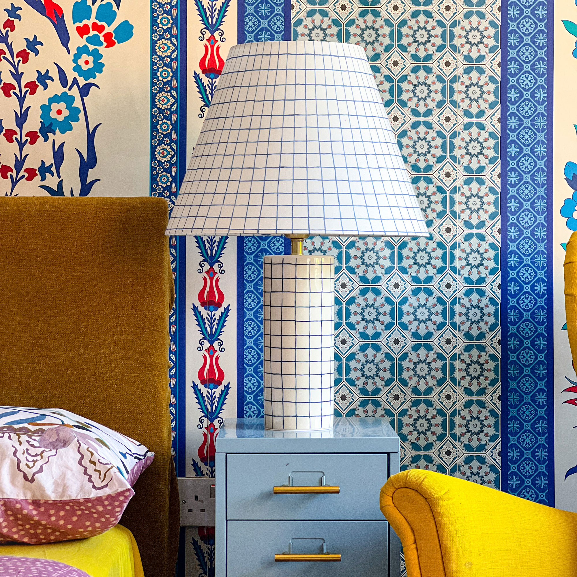 White lamp with blue graphic square pattern on base and shade, on light blue cabinet in front of wall with colourful, patterned wallpaper