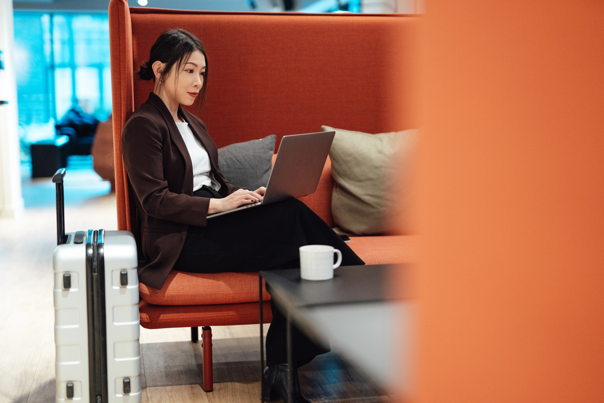 Woman sits with coffee and suitcase beside her as she works in airport lounge.