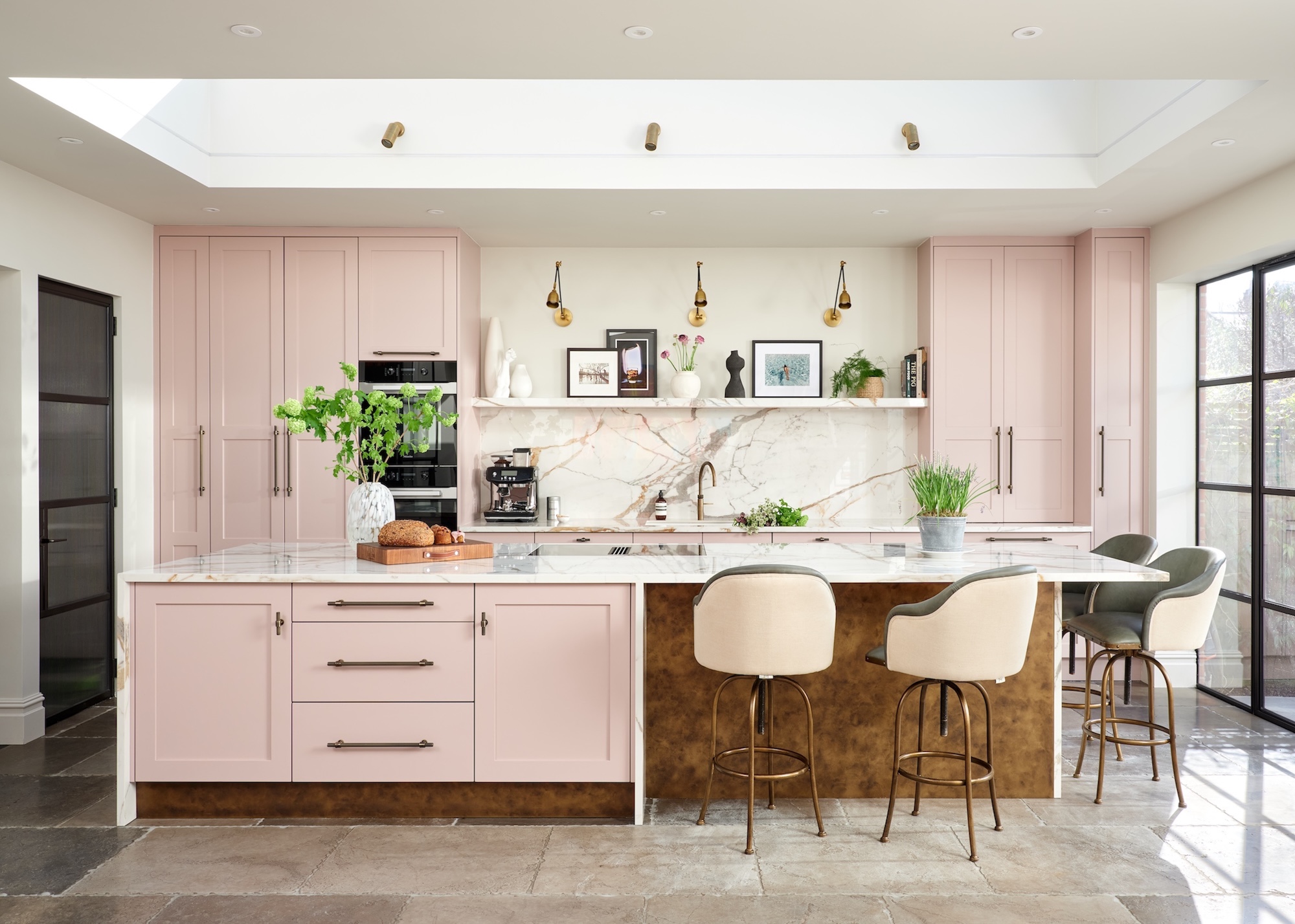 A modern kitchen with a view of the large kitchen island and pink cupboards with marble worktops and back splash. There is a large skylight above the island and cream bar stools around.