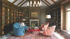 A grand, wood-paneled home library with floor-to-ceiling bookshelves, a vaulted beamed ceiling, a large tiered chandelier, and a plush blue sofa facing patterned coral armchairs over a Persian rug
