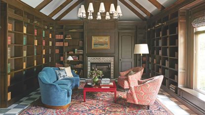 A grand, wood-paneled home library with floor-to-ceiling bookshelves, a vaulted beamed ceiling, a large tiered chandelier, and a plush blue sofa facing patterned coral armchairs over a Persian rug