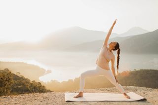 Athletic lady stretching and warming up on yoga mat. Fantastic scenery on background. Hills in mist and sea bay. Spiritual harmony with nature, mental health, active and healthy lifestyle. Life balance and vitality.